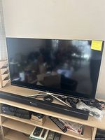 Samsung TV on stand with sound bar underneath and accessories around. TV is flat-screen, approx 35-inch size, screen off, note on top right corner.