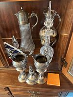 Photo showing silver plated hot water urn, coffee carafe with warmer, vintage wine carafe with crystal insert, two crystal salt and pepper shakers and multiple silver plated decorative containers with cherub figures