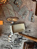 Photo showing multiple pieces of silver plated silverware spread on carpet, some stored in fabric rolls, a white container, and a wooden item handle visible