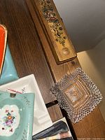 Photo showing part of the ashtray collection including blue floral ashtray, clear glass textured square ashtray, and a wooden item with decorative painting.