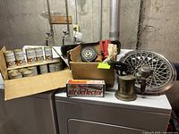 Wide shot of lot including six vintage Quicksilver Formula 50 motor oil cans, cardboard box with assorted items, antique brass blow torch, air deflector box, and power by Ford hubcap on top of white appliance.