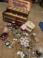Wide view of the lot showing the gold-tone Buxton jewelry box, assorted jewelry, pins, earrings, beads, and lockets spread on carpet.