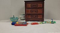 Photo showing wooden three-drawer chest, porcelain jar with lid, green ceramic bowl, and decorative tassels, all arranged on a white cloth.