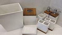 Photo of bathroom accessory set including waste bin, tissue box cover, soap dispensers, toothbrush holders, and soap dish against a cardboard backdrop