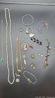 Varied costume jewelry displayed on a dark surface including necklaces, bracelets, rings, earrings and a brooch.