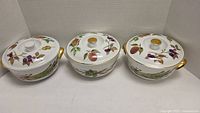 Three white ceramic casserole dishes with lids, each with colorful fruit and vegetable painted designs and gold accents on handles and lid knobs