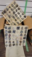 Two labeled display cards each holding multiple small rock specimens in individual compartments, showing a diverse mineral collection with names.