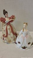 Photo showing two lady figurines: Lennox First Waltz in a cream and pink floral gown and Royal Doulton Old Country Rose in white dress with floral accents.