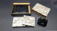 Photo showing the men's black leather Mundi wallet in original box, three mother of pearl vintage compacts stacked, and small black velvet purse ring box