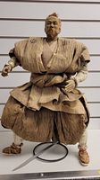 Paper mache samurai figurine front view in traditional attire, posed on a display stand with no sword visible.