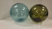 Two large glass floats side by side on white backdrop, one light blue and one olive green