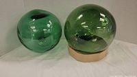 Two large round vintage glass fishing floats placed side by side on a white surface, one is dark green and the other light green.