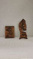Front view of two indigenous wood carvings - one rectangular plaque and one tall carving shaped like a bird figure.