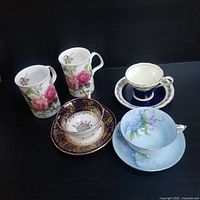 Group image showing four cups with floral designs: two white coffee cups with pink roses, one blue and gold ornate tea cup with saucer, and one light blue hydrangea teacup with saucer.