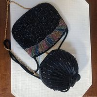 Photo shows two black beaded purses on white surface, one rectangular with multicolor leaf bead detail and gold chain, one round with drawstring closure