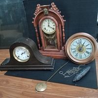 Photo showing all three clocks arranged together along with clock parts including a pendulum, chain, and pinecone weight.