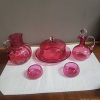 Full set of cranberry glassware including pitcher, covered butter dish with plate, decanter with stopper, and two small bowls displayed on white cloth on wooden surface.