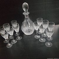 Full view of etched glass decanter with stopper and 10 matching etched wine glasses arranged around it.