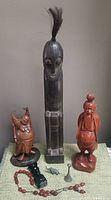 Photo shows three ethnic hand carved statues and several small artifacts on a surface against a beige wall. The tallest statue is dark wood with horsehair tuft, flanked by a reddish brown and a light brown statue. Several small metal artifacts and bead necklaces lay in front.