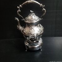 Front and side angle view of the hand chased silver tea pot on its heating stand, showing ornate floral engraving and curved handle.