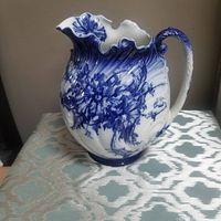 Front/view of large flow blue antique ceramic pitcher with distinct floral pattern and scalloped rim.