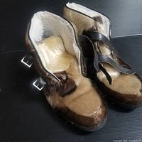 Pair of brown and tan fur winter boots with white plush lining and black leather straps with buckles, shown from above and side angle.