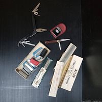 Photo showing multi-tool open with various blades and pliers, Ronson lighter in red leather case, two folding knives, nylon dress combs with packaging, and assorted small items on dark background.