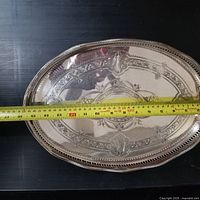 Top view of oval silver-toned tray with measuring tape showing approximately 13 inch length and visible embossed center design.