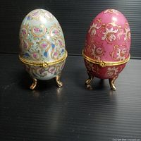 Front view of two porcelain egg trinket boxes, showing detailed floral patterns and gold metal feet.