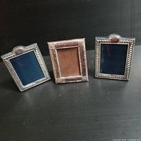 Three rectangular silver photo frames standing side by side on a black surface, showing front view with decorative borders and dark blue or brown backing material.