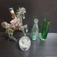 Photo showing four glass items: horse-shaped decanter, rounded etched glass piece, small bottle, and tall green vase.