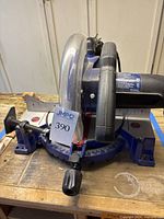 Front and side view of the compound miter saw showing blue base with angle measurement scale, black handle and clear blade guard, placed on wooden surface.
