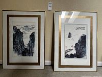 Front view of two framed Asian art prints side by side leaning against a wall showing black ink landscape scenes with gold matting and slim metallic frames