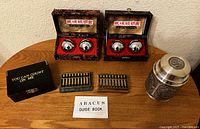 Two sets of polished metal Chinese iron balls housed in decorative cushioned boxes, two mini abaci including one detached from its case, an abacus guide booklet, a steel piggy bank with lid removed, all arranged on a wooden surface.