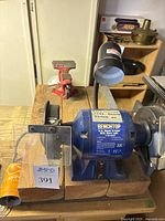 Full view of a blue benchtop grinder mounted on a wooden table with a red clamp visible behind it.