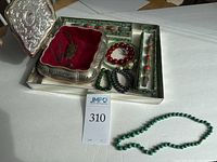 Photo showing the pewter jewelry box with red velvet interior containing carved stone pendant on black cord, red carnelian beaded bracelet, green malachite and shiny hematite bracelets, and green jade necklace beside it.