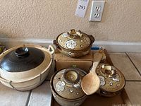 Four lidded clay casseroles displayed together on floor, three painted with flowers, one larger plain beige pot with black lid secured by wire handle. Large wooden spoon rests on top of smaller pots. Small painted bottle visible beside pots.