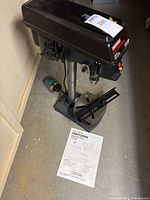 Full view of the drill press showing base, column, table, and head along with the instruction manual on the floor.