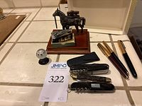 Photo of entire lot including horse lighter, Flying Swallow lighter with box, pens, pocket knives, and black case on tiled surface.