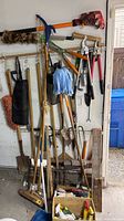 Photo of long-handle gardening tools and gloves hanging on wall rack, black rubber apron visible