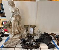 Photo shows classical-style statue of a woman carrying a basket, two candlesticks, small planter, and decorative black fan-shaped sconce grouped together on a tile surface.