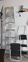 Full view of the 7-foot aluminum ladder, two small step stools with black rubber treads, and the Fiskars branch lopper hanging on the wall.