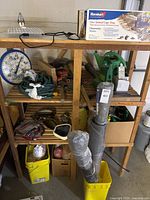 View of wooden shelving unit holding temperature gauge, green watering cans, live animal cage trap box, coiled hoses, and assorted gardening supplies in containers and boxes.