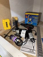 Overview of all items: Craftsman hammer drill, two yellow DeWalt bit containers, portable light with box, and carpenter's square, placed on wooden surface.