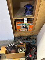 Photo showing a blue Benchtop Pro palm grip sander placed on a wooden shelf with power cord wrapped. Below shelf, original box and tool visible.