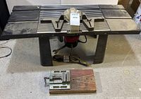 Overall view of Craftsman router table showing metal table top with adjustable fences and router motor below, power cord coiled on ground. Clamp mounted on wooden base placed in front.