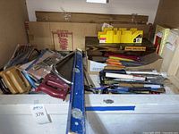 Wide shot of multiple hand saws, a long blue spirit level, wood carving tool set in wooden box, and other assorted woodworking tools piled in a container.