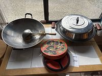 Overview of two large steel woks, one metal lid, one large scoop, round covered serving dish placed on a surface near window.