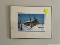 Framed giclee print of a snow-covered sugar shack with bare trees and blue sky, mounted on a white mat and framed in silver metal frame.