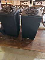 Pair of black rectangular speakers with speaker wires coiled on top, placed on wooden surface.